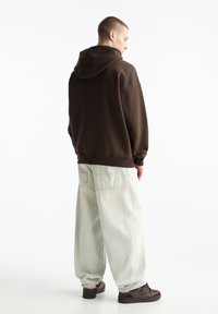 Braune oversized Kapuzenjacke mit Kordelzug, kombiniert mit hellgrünen, lockeren Jeans. Dunkle Schuhe sind sichtbar. Einfacher Hintergrund.
