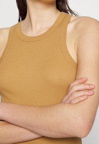 Femme portant un haut sans manches côtelé couleur moutarde, bras croisés sur la poitrine, sur fond uni.