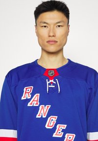 Tricou de hochei albastru cu "RANGERS" scris cu litere albe și roșii. Are un guler cu șnur și logo-ul NHL pe un accent roșu la gât.