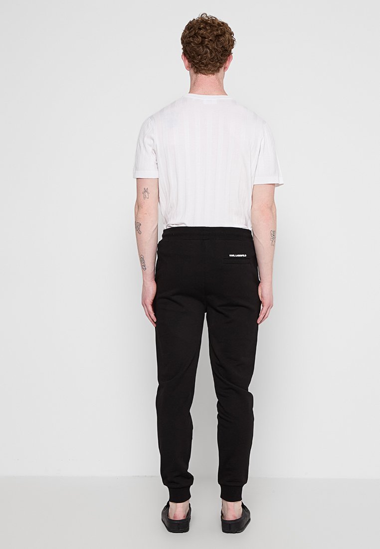 Schwarze Sweatpants mit schmaler Passform und elastischem Bund, kombiniert mit einem weißen, gerippten T-Shirt. Dazu werden schwarze Slipper getragen.