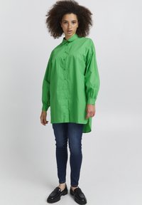 Green oversized button-up shirt with long sleeves and a high-low hem, paired with dark skinny jeans and black shoes. Smooth texture.