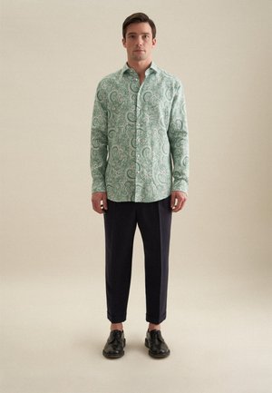 Camicia button-up verde a motivo paisley, realizzata in tessuto leggero, abbinata a pantaloni scuri blu navy e scarpe nere stringate.