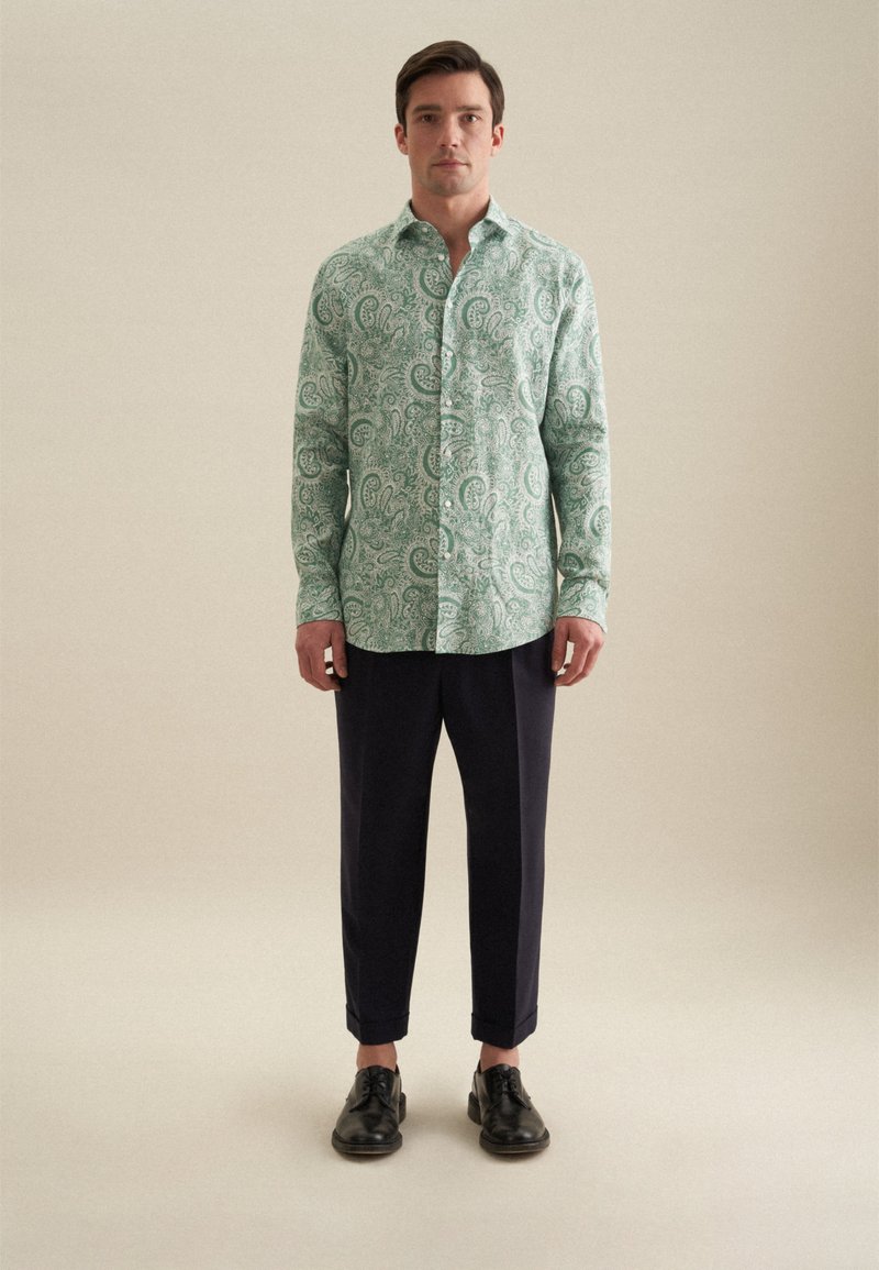Camicia button-up verde a motivo paisley, realizzata in tessuto leggero, abbinata a pantaloni scuri blu navy e scarpe nere stringate.