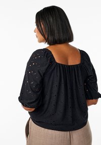 Blusa nera in tessuto con dettagli a occhiello, maniche a sbuffo e scollatura quadrata, indossata con pantaloni marroni chiari.