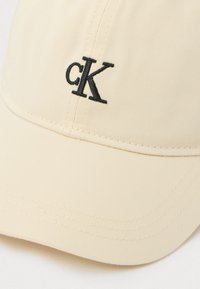 Bežs cepure izgatavota no teksturēta auduma ar izliekta mala, ar melnu izšuvumu 'cK' logotipu priekšā.