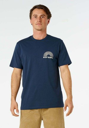 Uomo che indossa una t-shirt Rip Curl blu navy e pantaloni marrone chiaro, in piedi davanti a uno sfondo bianco semplice.