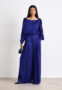Ensemble en satin violet comprenant un haut à manches longues et un bas à jambes larges, avec des rayures verticales. Les accessoires incluent un petit sac noir structuré.
