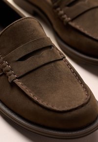 Mocassins penny en daim marron avec couture détaillée et bout arrondi, montrés en gros plan sur une surface claire.