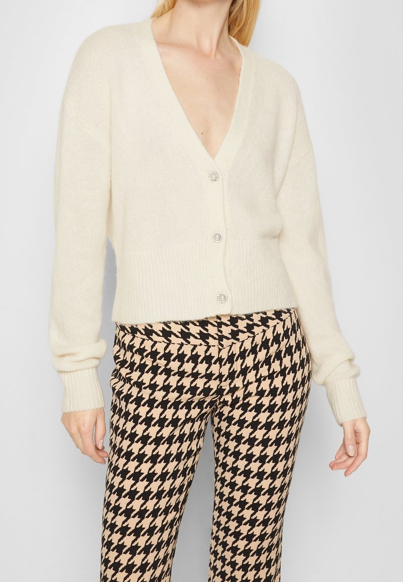 Cardigan color crema con scollo a V, tessuto morbido, orlo e polsini a coste, con bottoni decorativi. Abbinato a pantaloni in tessuto a scacchi houndstooth.