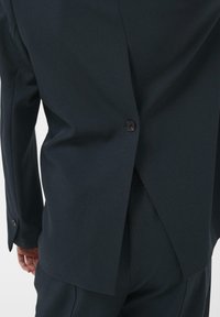 Tengerészkék öltönykabát, egyetlen gombbal, testre szabott szabásvonalakkal, sima textúrával és hátsó hasítékkal a mozgékonyság érdekében.