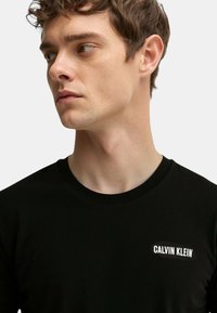 Giovane uomo con capelli corti castani che indossa una maglietta nera di Calvin Klein, mentre guarda a sinistra su uno sfondo chiaro e semplice.