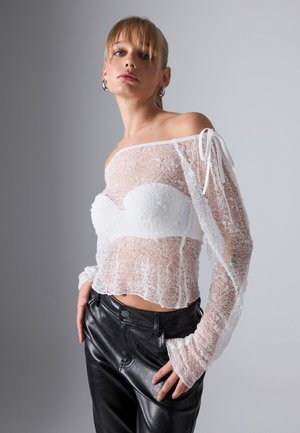 Hvit off-shoulder blonde topp med lange ermer, med blomsterdesign, gjennomsiktig tekstur og båndknuter på skuldrene, parret med svarte skinnbukser.