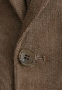 Le tissu en velours côtelé marron est orné d'un grand bouton rond avec quatre trous. Le bouton est de couleur claire et a une texture lisse.