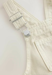 Beige striped overalls featuring a silver metal buckle and clip, with visible stitching details and a front pocket. Soft fabric texture.