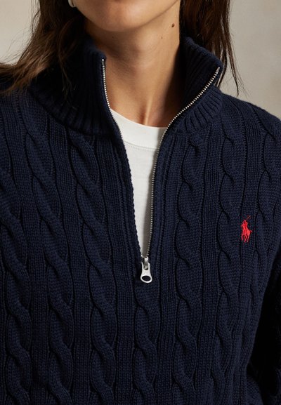 Jersey azul marino de punto trenzado con medio cierre y cremallera plateada, con logotipo rojo bordado, llevado sobre una camiseta blanca de cuello redondo.