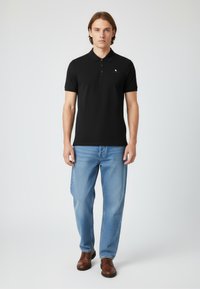 Svart polotröja med en två-knapps knappslå, kortärmad, och en liten logotyp på bröstet, ihop med ljusblå jeans och bruna skor.