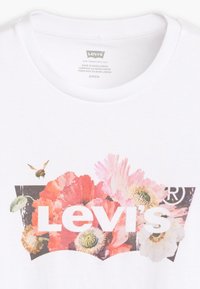 Vit bomull t-shirt med en färgglad blommig grafik och "Levi's" logotyp i fet vit text mot en mörk bakgrund.