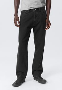 Black denim jeans with a straight fit, five-pocket design, and subtle stitching details, paired with black sneakers and a gray shirt.