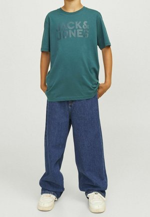 Dítě v zeleném tričku "Jack & Jones", s volnými modrými džínami a bílými teniskami, stojící s rukama v kapse.