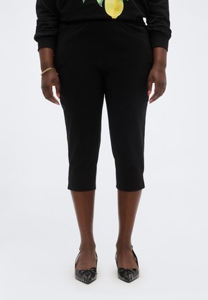 Personne portant un pantalon noir court et des escarpins noirs à bout pointu, debout devant un fond blanc uni.