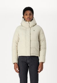 Beige prešívaná bunda s vysokým golierom, predným zipsom a prešívaným dizajnom. Obsahuje malé logo na hrudi a elastické manžety.