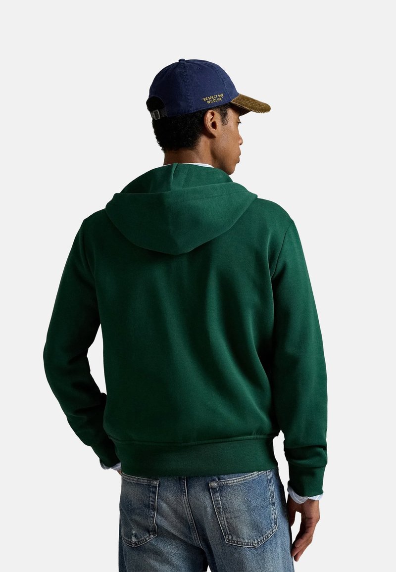 Sudadera verde con capucha hecha de una tela suave, que presenta un bolsillo tipo canguro y puños acanalados. Se lleva con unos jeans desgastados de color azul claro y una gorra azul marino.