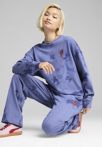 Felpa con tie-dye viola e pantaloni abbinati, con dettagli di sole ricamati. Il modello indossa sneakers rosa e rosse, mostrando un fit rilassato.