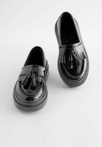 Sorte patentlæder loafers har en frynset detalje og en flad, struktureret gummisål. Designet inkluderer dekorativ syning langs kanterne.