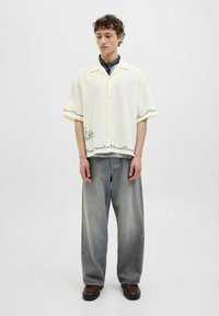 Chemise jaune à manches courtes avec broderie florale verte à l'ourlet, associée à un pantalon en denim délavé à coupe décontractée et des chaussures marron.