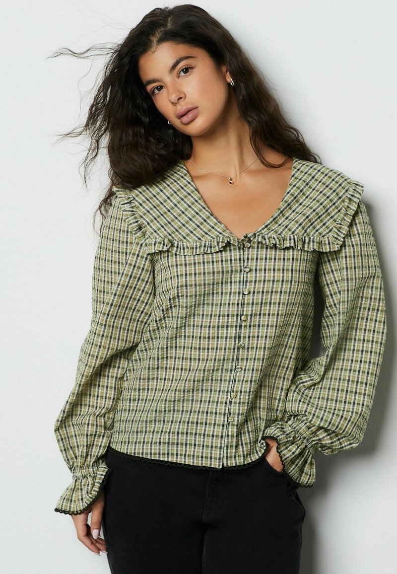 Blouse verte à carreaux avec col à volants ; manches longues avec poignets froncés ; boutonnage sur le devant ; tissu léger.