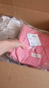 Ροζ ρούχο σε διαφανή πλαστική συσκευασία, μερικώς αποκαλυμμένο από ένα χαρτόνι. Ορατή ετικέτα με λεπτομέρειες προϊόντος προσαρτημένη.