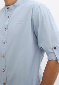 Camicetta azzurro chiaro con colletto alla mandarino, bottoni in legno, maniche arrotolate e un piccolo logo ricamato di un cervo sul petto.
