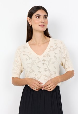 Frau mit langen dunklen Haaren trägt ein cremefarbenes kurzärmeliges V-Ausschnitt-Stricktop mit Blumenmuster und einen schwarzen Rock, schaut leicht zur Seite.