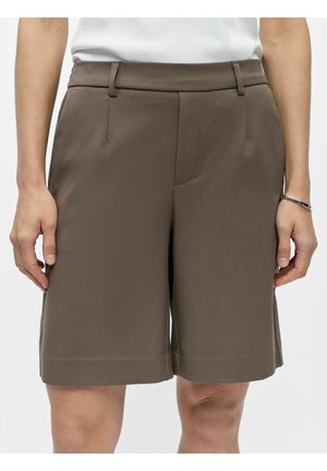 Mittelange braune, maßgeschneiderte Shorts mit Gürtelschlaufen und Seitentaschen, getragen von einer Person mit weißem Oberteil und einem Armband am rechten Handgelenk.