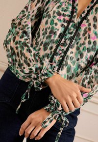 Caura blūze ar zaļu, rozā un melnu leoparda rakstu; piedāvā savilktas piedurknes un melnu pogu apdari ap kaklu.