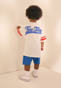 Vitt T-shirt med rödrandiga ärmar och blå "Tom och Jerry"-grafik. Matchas med blå shorts och vita sneakers med röda detaljer.