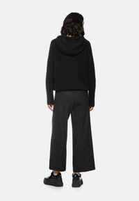 Schwarzer gestrickter Kapuzenpullover mit boxy Schnitt, ausgestattet mit einer Fronttasche und einer samtigen Textur. Kombiniert mit locker sitzenden schwarzen Hosen und Sneakers.