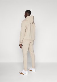 Beige hettegenser og matchende joggebukser laget av mykt stoff, med et logo-merke på ryggen, sammen med hvite og brune joggesko.