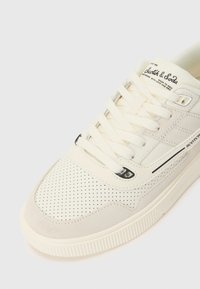 Cream-colored sneaker with perforated texture, suede accents, and a rubber sole. Features flat laces and branding on the tongue.