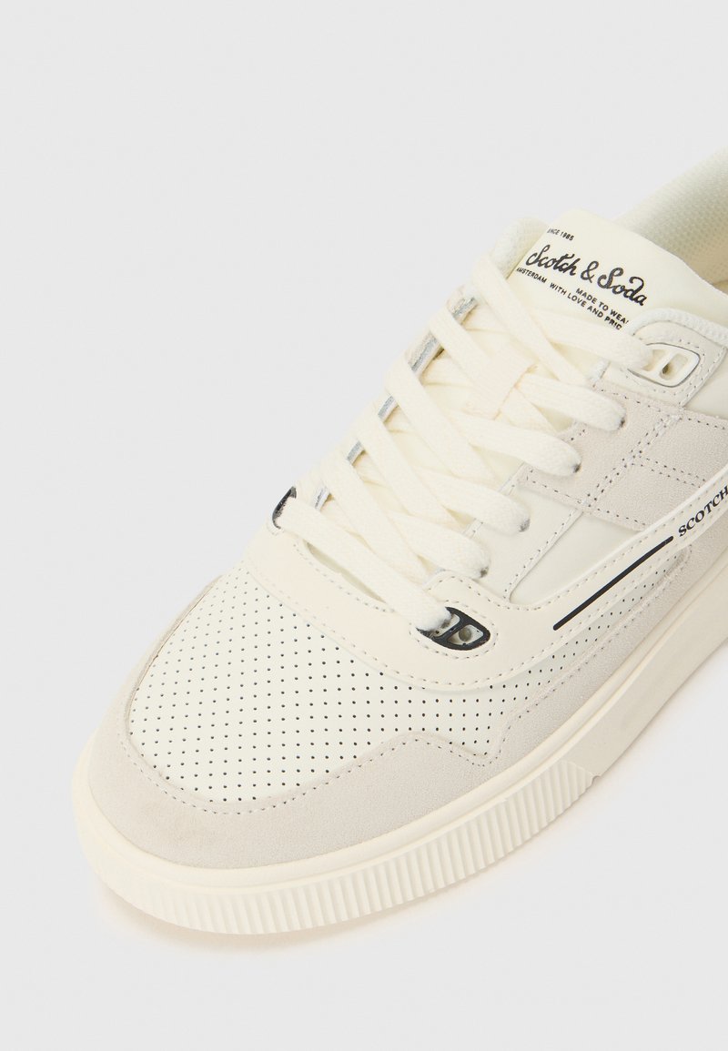 Crèmekleurige sneaker met geperforeerde structuur, suede accenten en een rubberen zool. Beschikt over platte veters en merklogo op de tong.