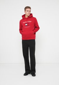 Red hoodie with front pocket and drawstring, featuring white "Tommy Hilfiger" logo and tricolor flag design. Black trousers and shoes.