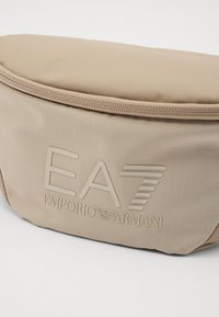 Beige nylon deréktáska sima felülettel, cipzáras záródással és a „EA7 Emporio Armani” dombornyomott logójával az elején.