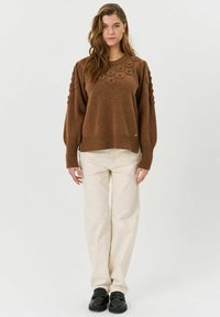 Brauner gestrickter Pullover mit strukturiertem Blumenmuster auf den Schultern, gerippten Bündchen und Saum, kombiniert mit cremefarbenen Hosen und schwarzen Schuhen.