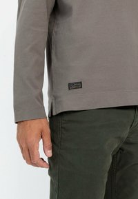 Grijze langemouwen shirt met een ronde halslijn, zijsplitten in de zoom en een label op de linkeronderkant. Draagt met donkergroene broek.