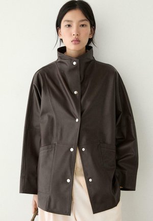 Kvinde iført en mørkebrun knapjakke med høj krave og løse ærmer, holder en lille brun taske mod en ensfarvet baggrund.