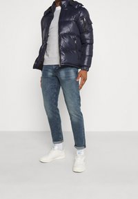 Chaqueta tipo puffer en color navy con textura brillante, bolsillos con cremallera y un cuello alto, combinada con una sudadera gris y unos jeans azules. Zapatillas blancas.