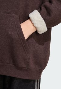 Felpa marrone con polsino interno in pile leggero grigio chiaro. Presenta una tasca frontale e un tessuto testurizzato con sfumature sottili.