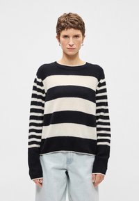 Suéter listrado com faixas horizontais pretas e creme, textura de malha, decote redondo e punhos canelados, combinado com calças de ganga claras.