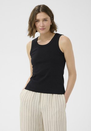Femme aux cheveux courts et bruns portant un haut noir sans manches et un pantalon blanc à fines rayures verticales, mains dans les poches, fond neutre.
