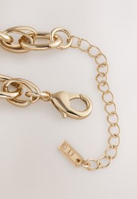 Gold chain accessory featuring large, interlocking links, a clasp, and a tag with a small accent stone. Smooth, polished metal finish.